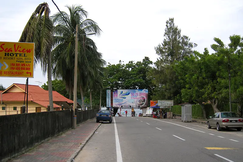 Street view leading to Pasir Bogak Beach on Pangkor Island with signage, palm trees, and hotels nearby.
