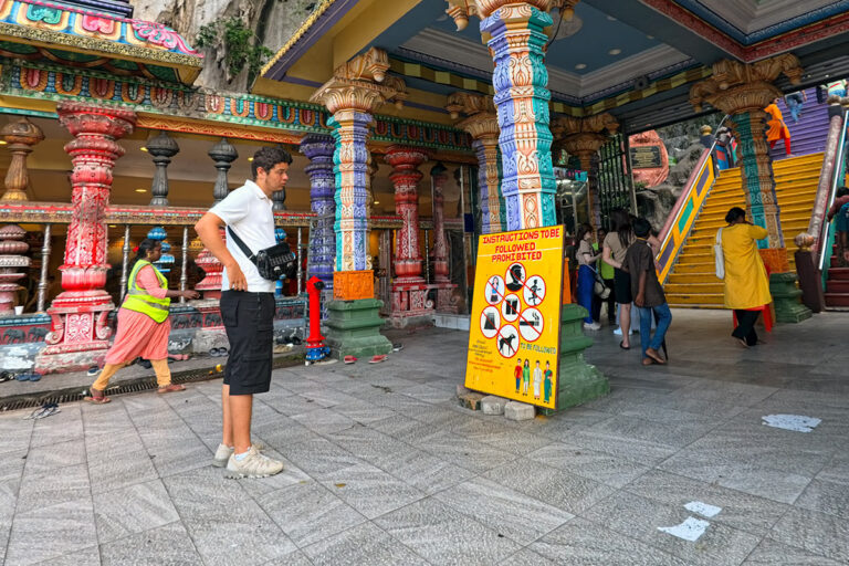 A tourist reading the dress code sign at Batu Caves entrance, with colorful temple pillars and stairs in the background.