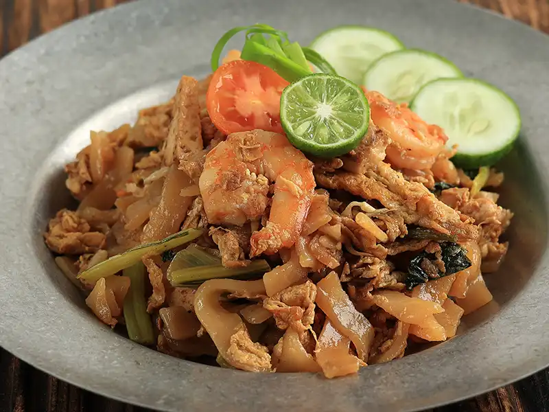 Char kway teow in Kuala Lumpur topped with prawns, egg and flat stir-fried noodles, served with lime and cucumber — a must-try Malaysian dish for first-time visitors.